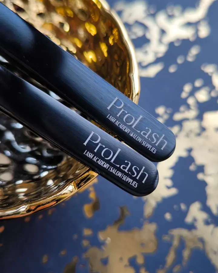 Two black ProLash tweezers resting on a gold bowl